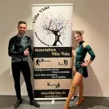 Un danseur et une danseuse en costumes de scène verts, de part et d’autre du roll-up Villa Vida.