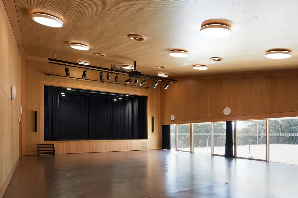 La grande salle des Utins à Echandens : parquet sombre, plafond en bois, scène aux rideaux noirs, baies vitrées.